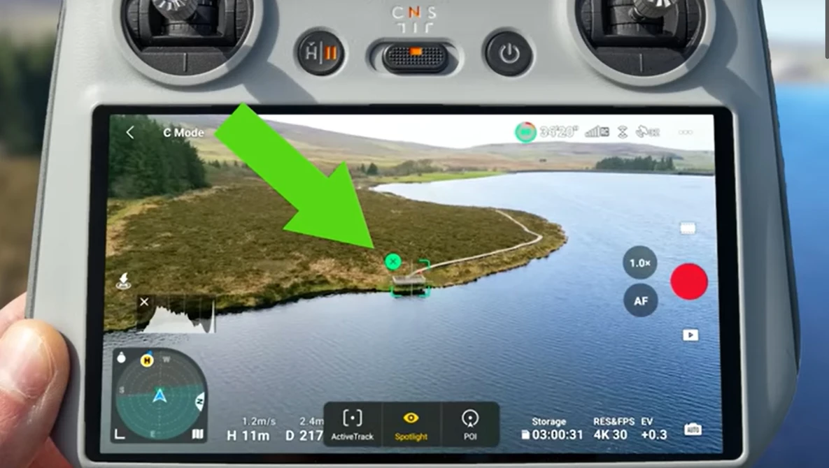 Screenshot of drone controller display showing aerial video feed of a curved shoreline with green arrow pointing to waypoint marker.