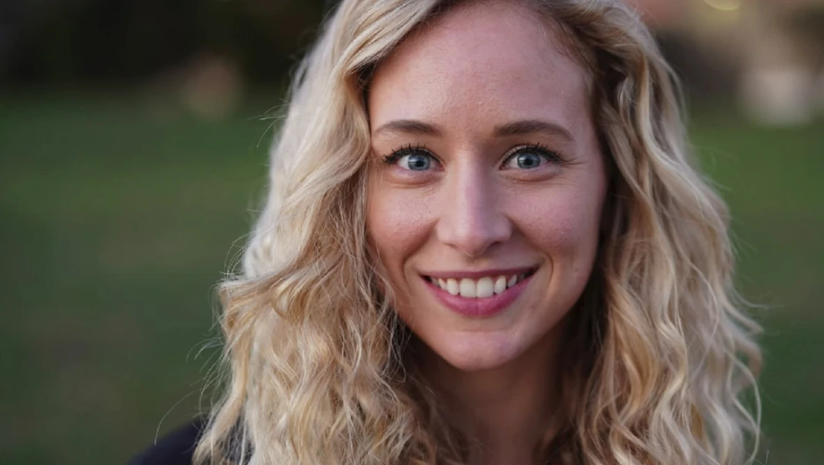 Woman with wavy blonde hair smiling at camera outdoors with blurred green background.