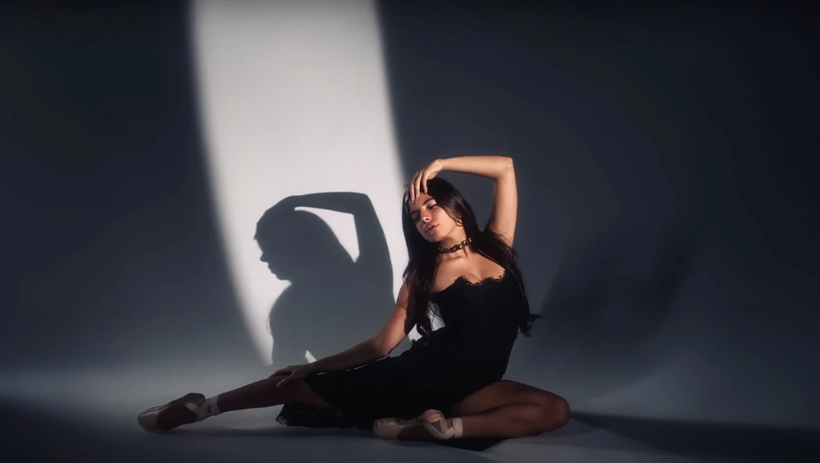Dancer in black performing a seated stretch pose with dramatic side lighting creating strong shadows.