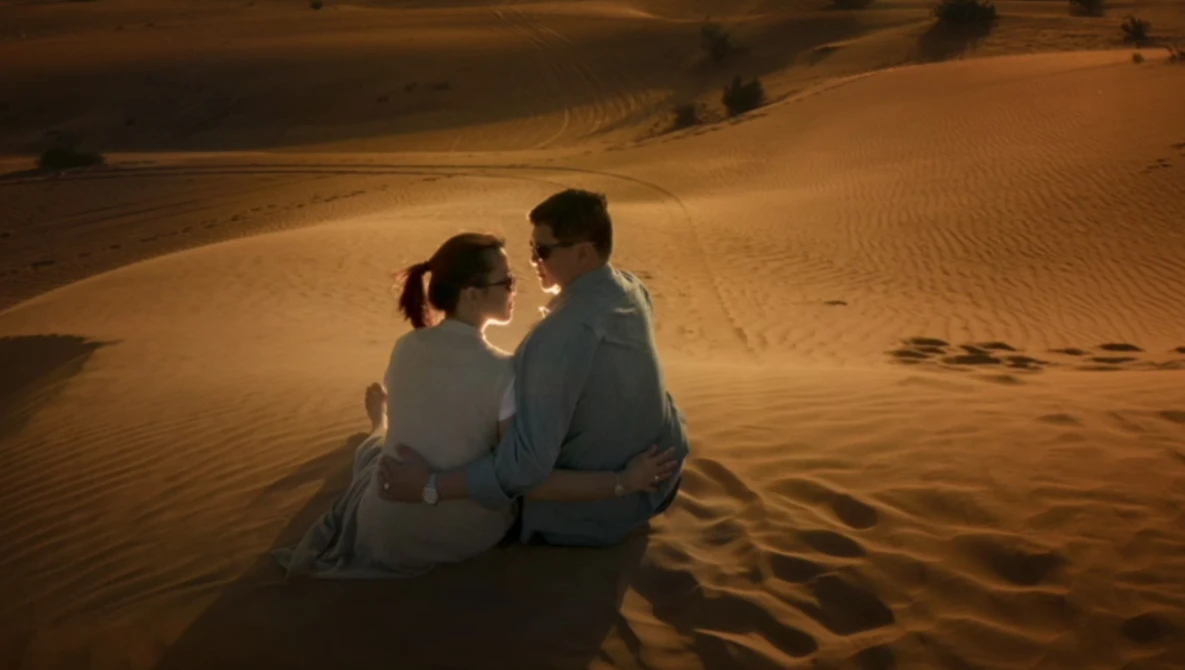 Couple sitting together on sand dunes at golden hour, facing each other in conversation.