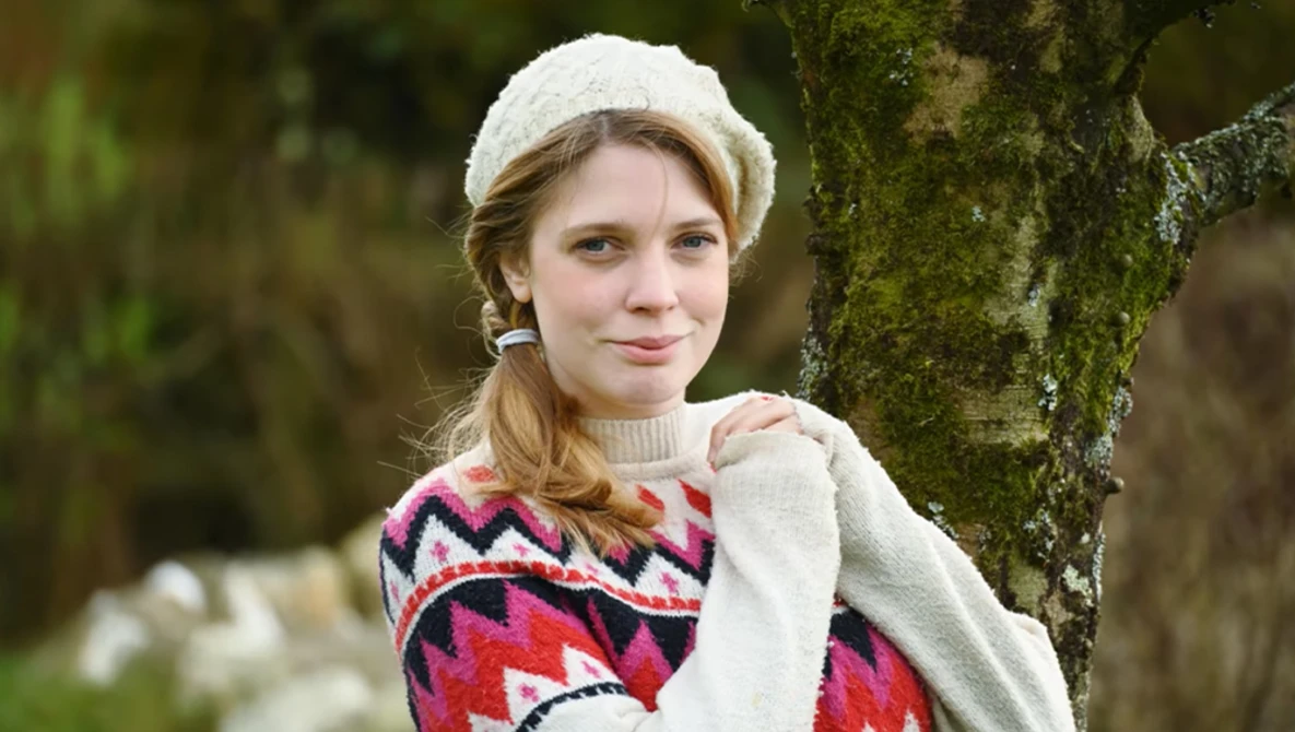 Young woman in cream knit beanie and red patterned sweater leaning against moss-covered tree trunk.