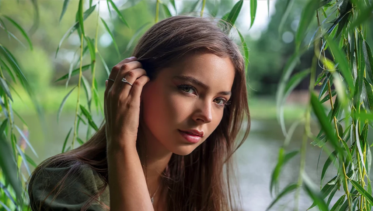 Young woman with long dark hair posing among hanging willow branches near water.