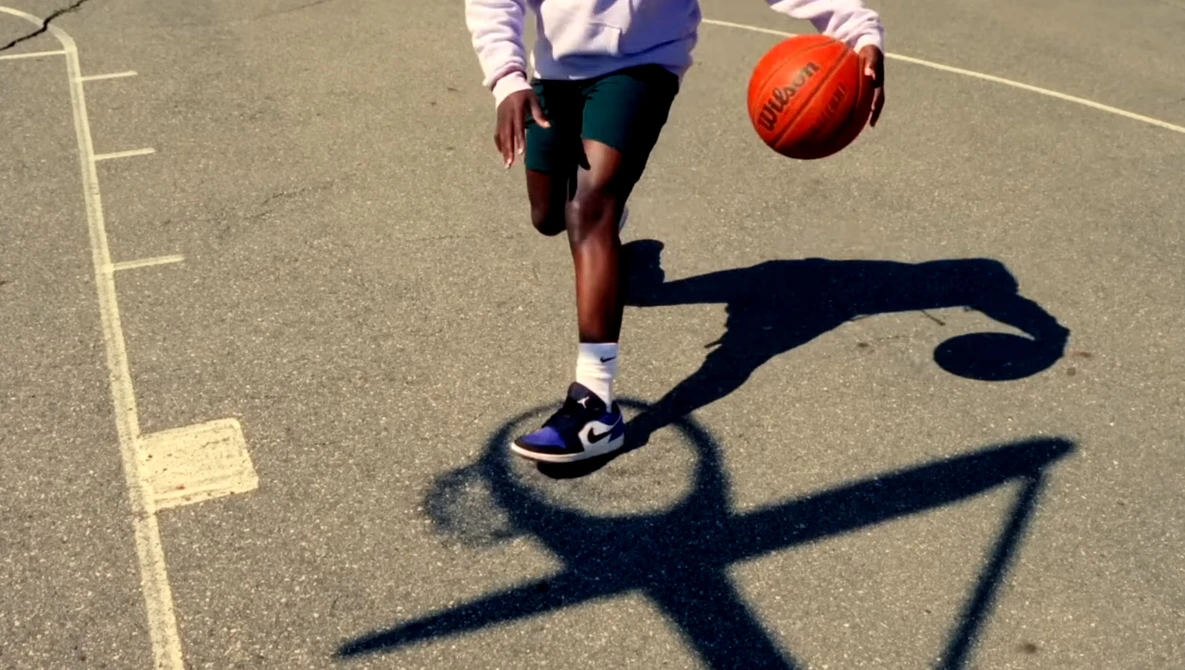 Player dribbling a basketball on an outdoor court with a sharp shadow cast by the sun.