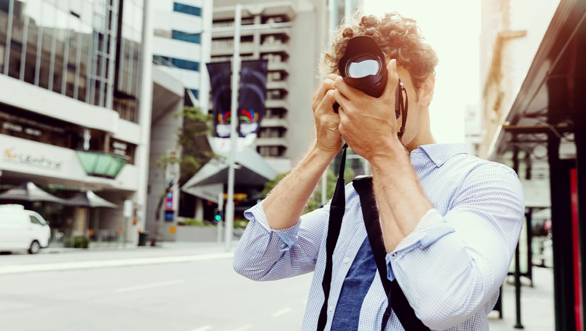 Man holding vintage camera up to his face on an urban street with tall buildings behind him.