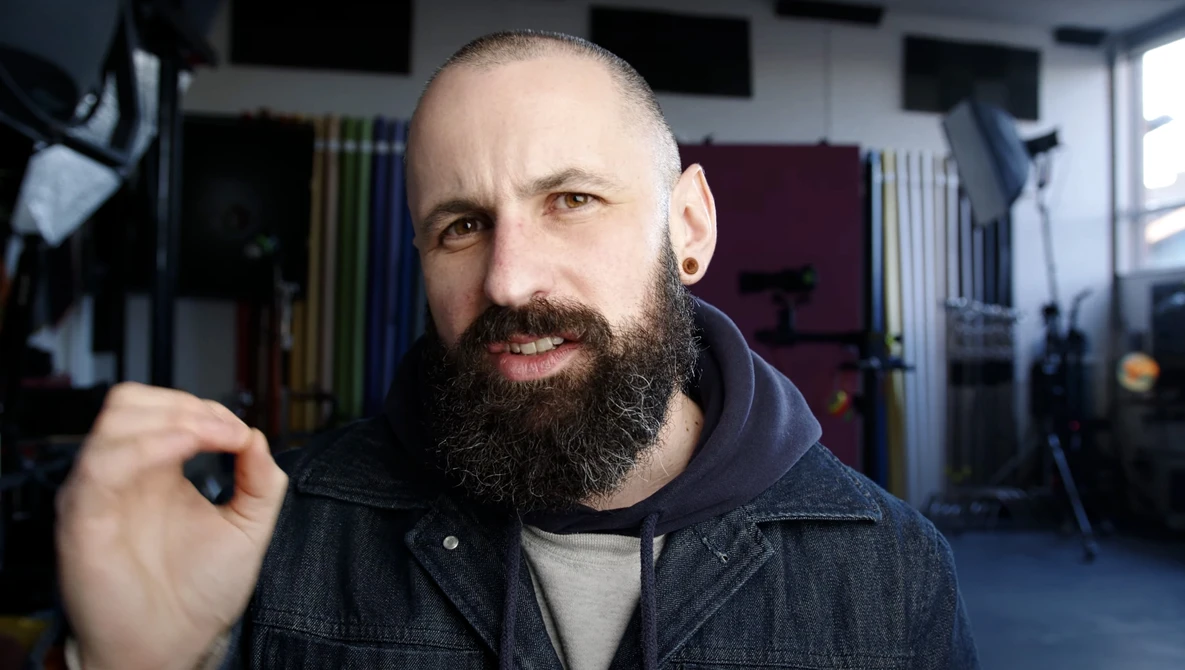 Bearded man with shaved head in denim jacket gestures while speaking in a studio setting.
