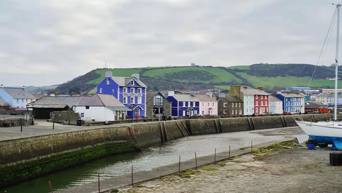 Colorful waterfront buildings reflected in calm harbor water, with sailboat and hillside backdrop.
