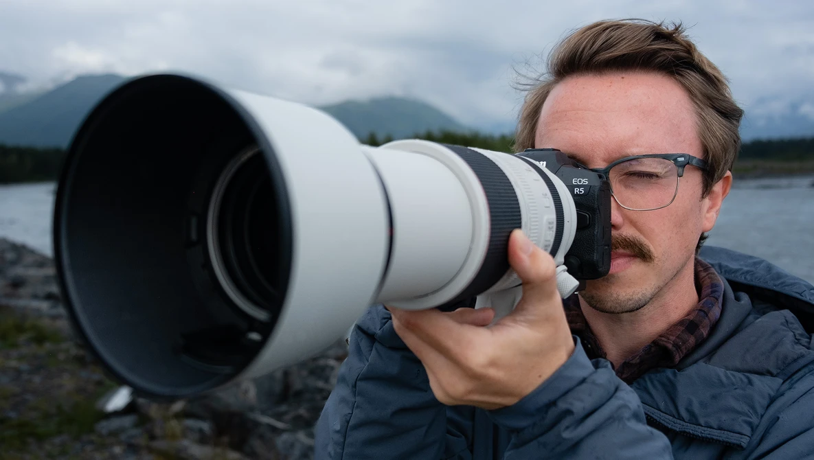 Photographer holding a white telephoto lens toward camera while wearing glasses and dark jacket by a lake.