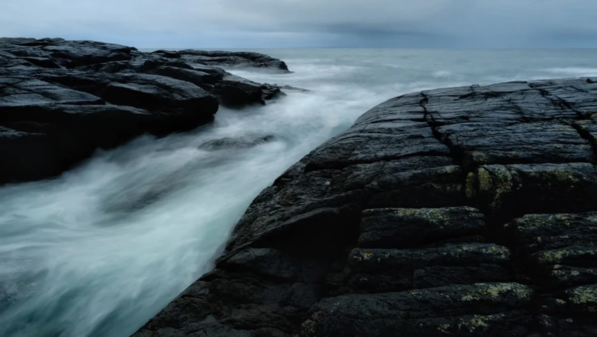 Turbulent seawater rushes between dark layered rock formations under an overcast sky.