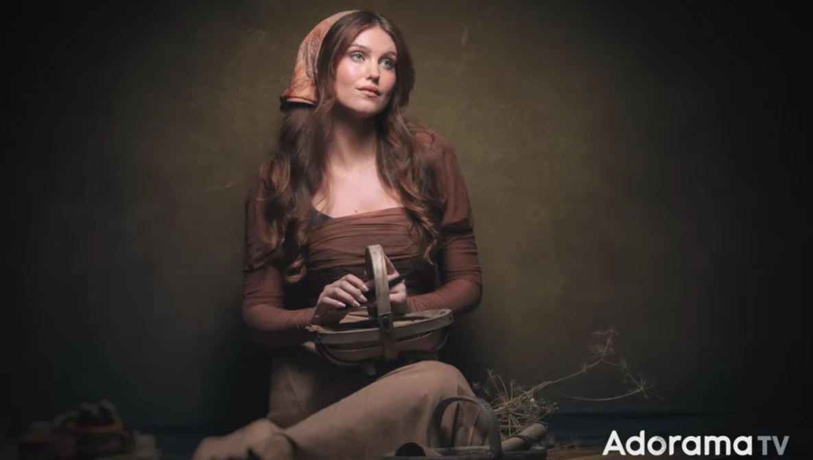 Woman in brown clothing holding a book, posed against a dark studio background with warm lighting.