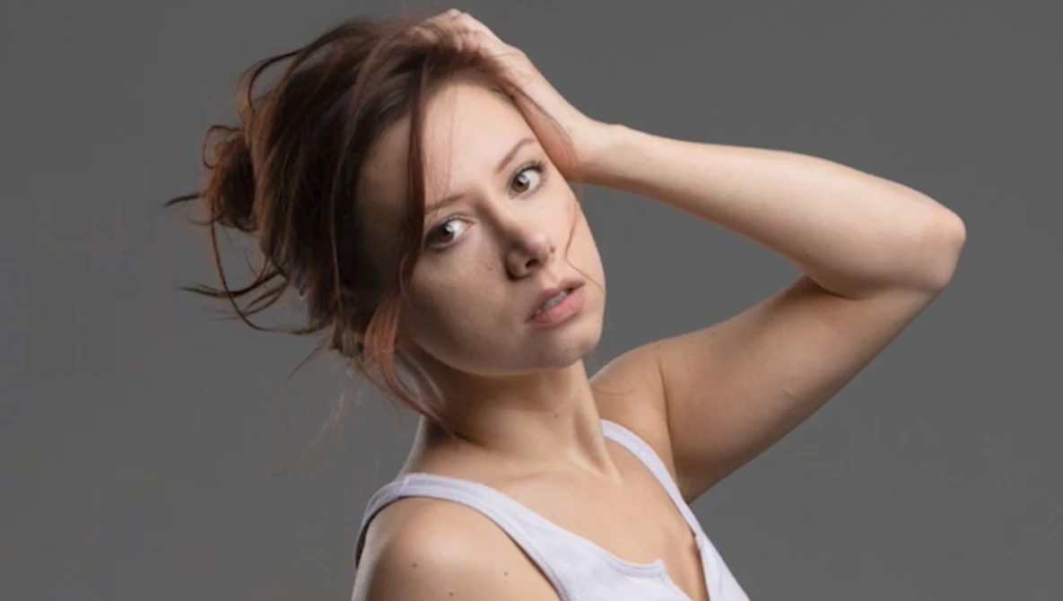 Woman with reddish-brown hair in casual white tank top against neutral gray background.