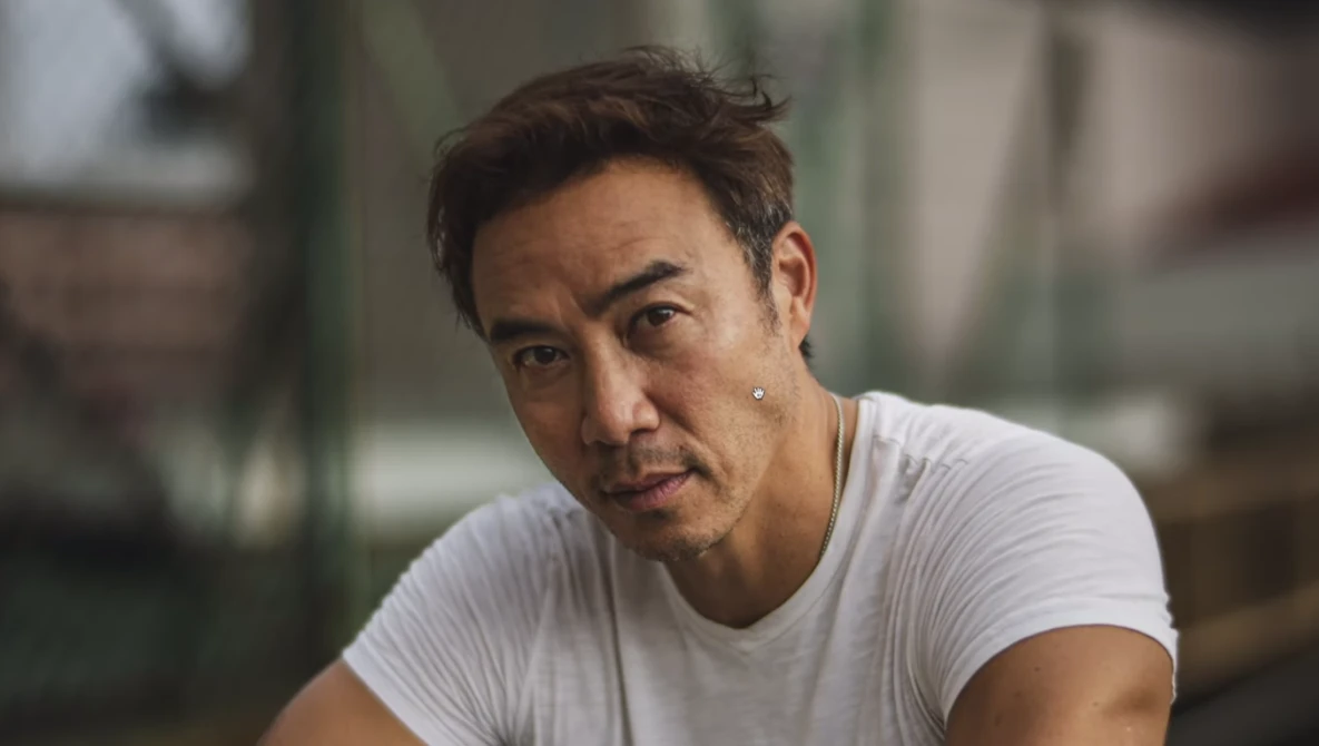 Man in white t-shirt looking directly at camera with serious expression.