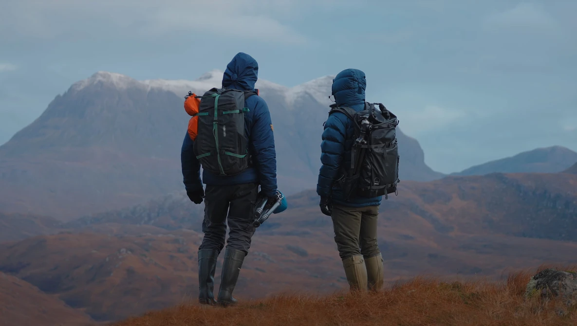 Two hikers with backpacks stand on a ridge overlooking mountainous terrain under partly cloudy skies.