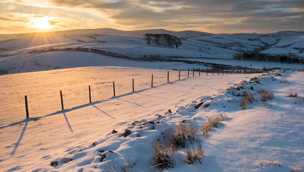Wooden fence line crossing snow-covered hillside at golden hour with distant forested ridge and mountains.