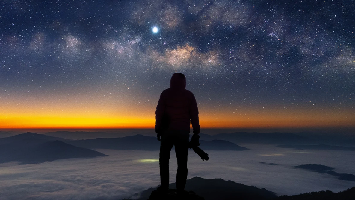 Silhouetted figure standing on a mountain peak gazing at the Milky Way and sunrise.