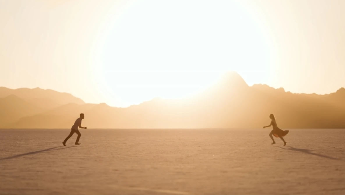 Two silhouetted figures in crouched positions on a vast sandy plain, backlit by a golden sunset behind distant mountains.