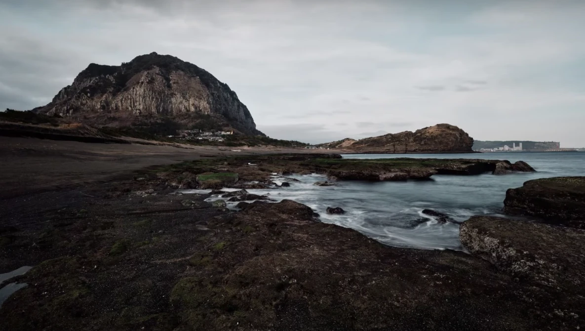 Rocky volcanic coastline with dark sand beach, prominent mountain peak, and calm ocean waters under overcast sky.