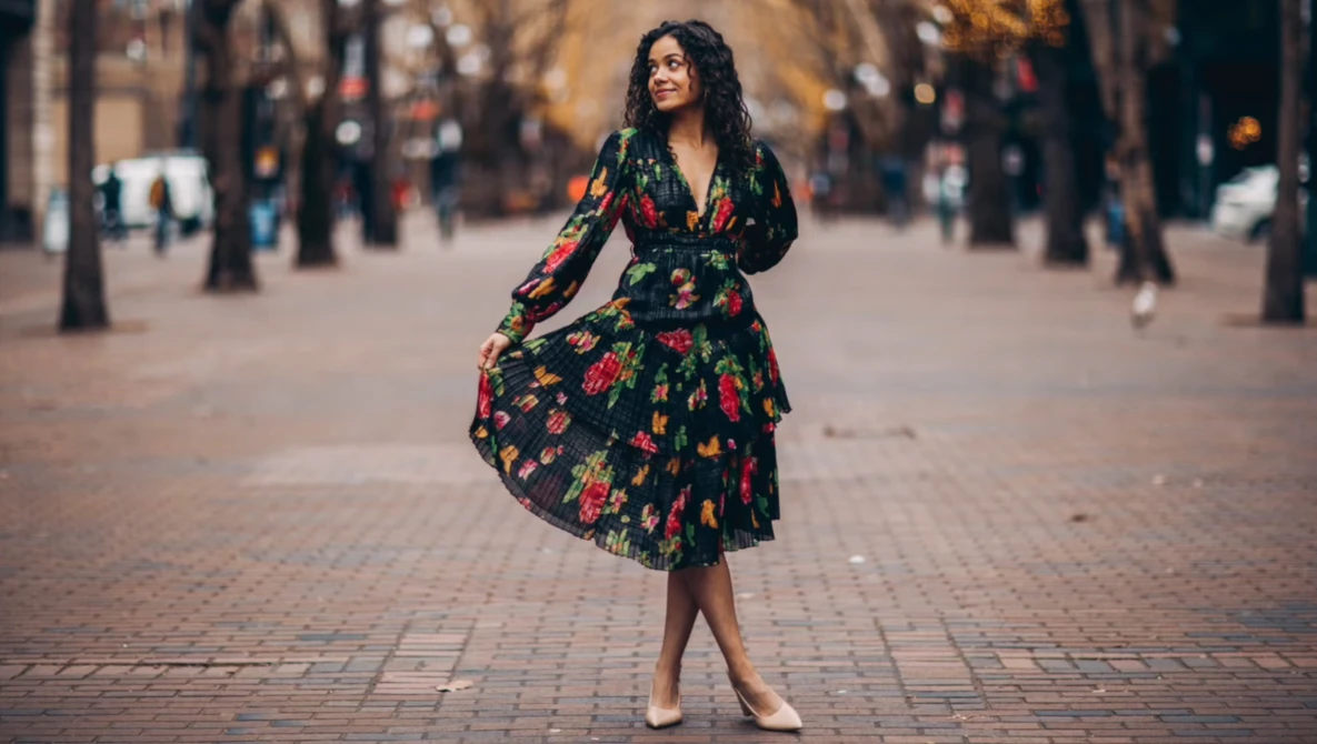 Woman in black floral dress standing on a brick plaza lined with trees.