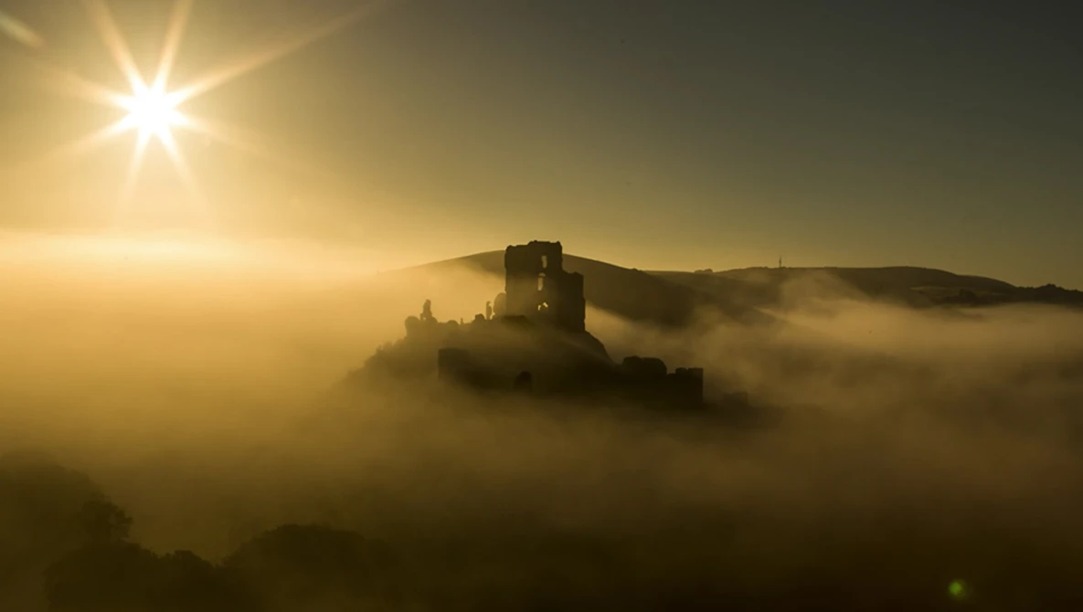 Medieval castle tower emerging from thick fog at sunrise with golden sunlight streaming overhead.