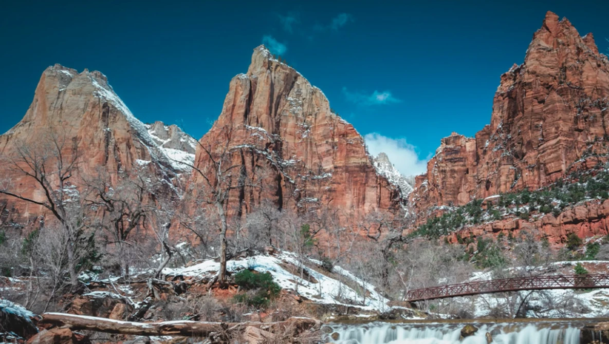 Red rock canyon with snow-capped peaks under clear blue sky and flowing water in foreground.