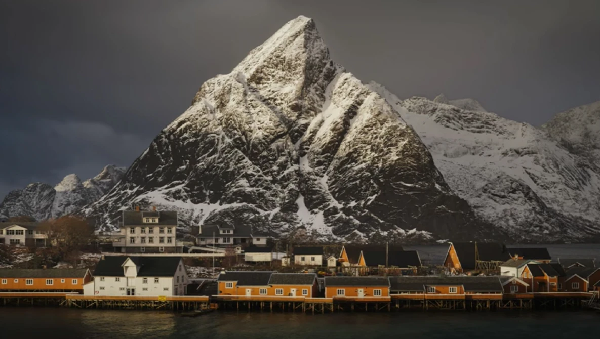 Snow-covered mountain peak rising behind a waterfront village with yellow and orange wooden buildings.