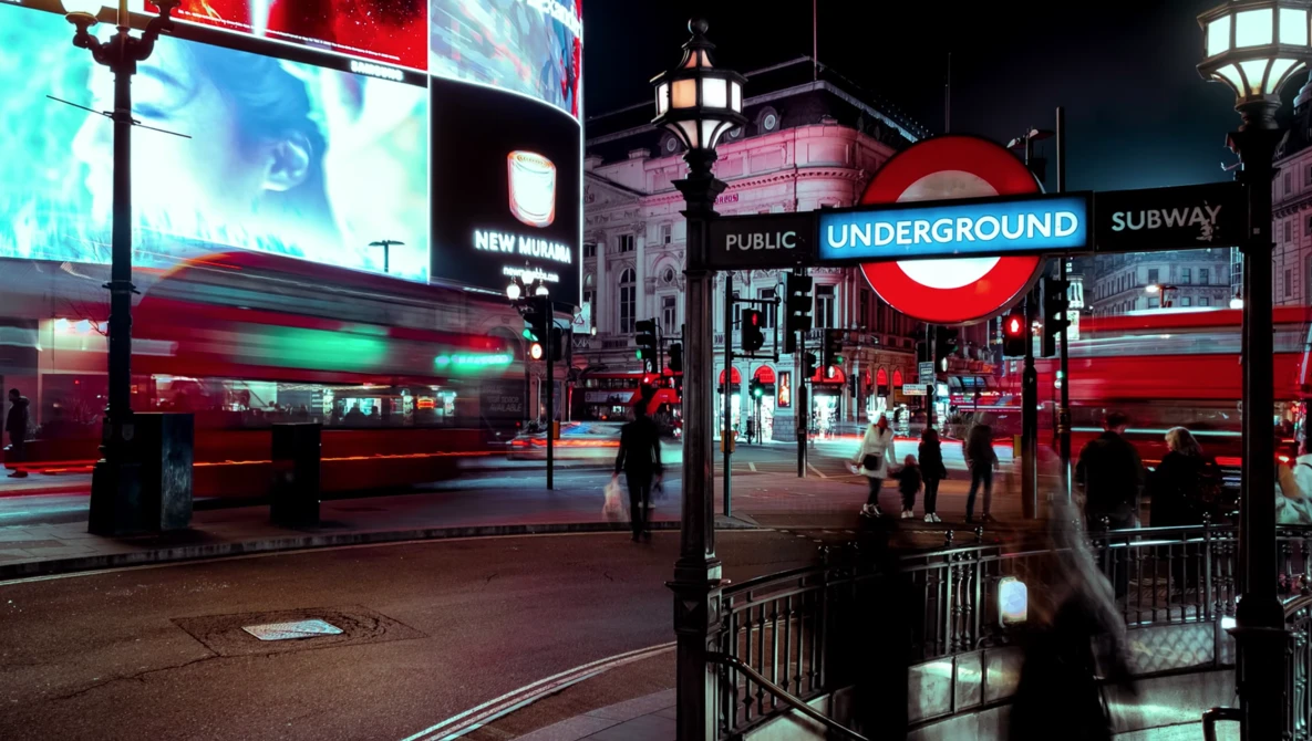 London Underground station entrance at night with neon signage and red double-decker bus in motion blur.