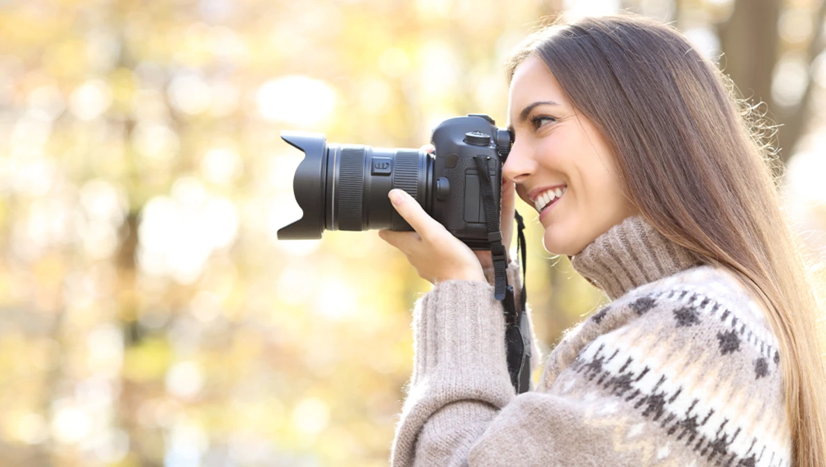 Woman holding a DSLR camera with telephoto lens, smiling while photographing outdoors.