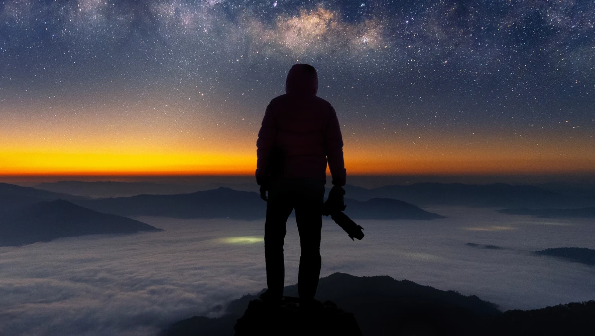 Silhouetted figure standing on a mountain peak overlooking a sea of clouds at sunrise, with the Milky Way visible in the night sky above.