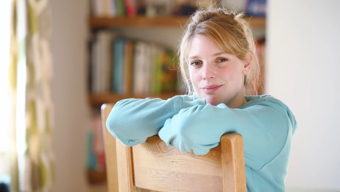 Young woman in light blue shirt leaning on wooden chair back, looking toward camera in home library setting.