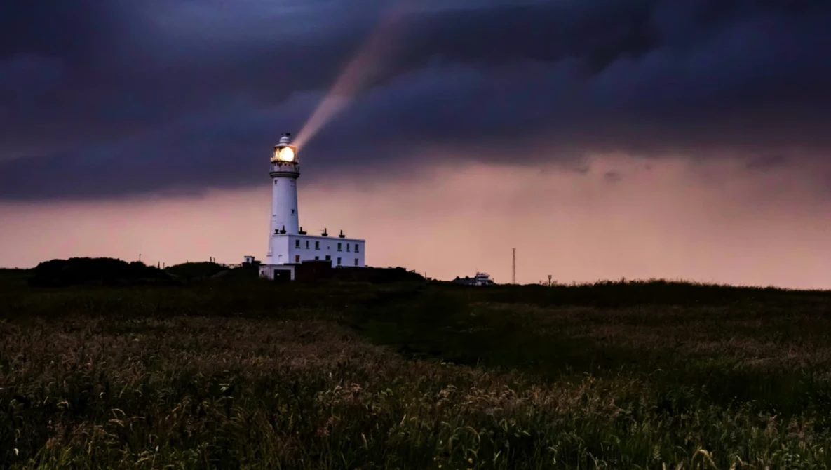 Illuminated lighthouse beam cutting through dramatic storm clouds at dusk over a grassy field.