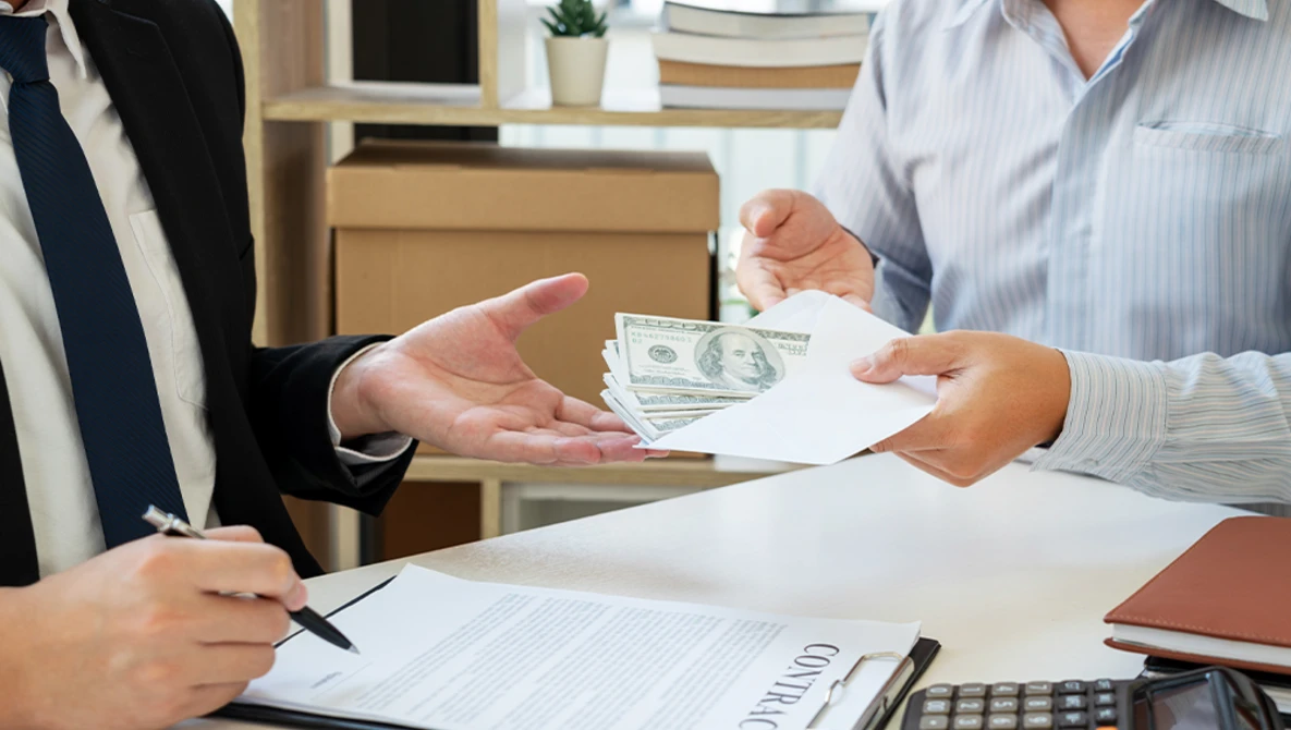 Two businesspeople exchanging cash and documents at a desk during a transaction.
