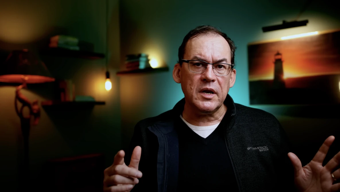 Man wearing glasses and dark jacket gestures while speaking in a studio setting with warm ambient lighting.