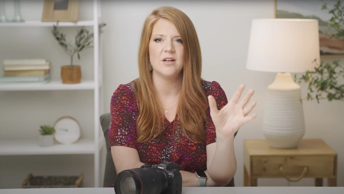 Woman with reddish-brown hair in burgundy patterned shirt gesturing while speaking in home interior setting.