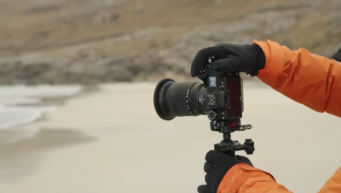 Photographer in orange jacket holding DSLR camera on tripod at beach during overcast conditions.