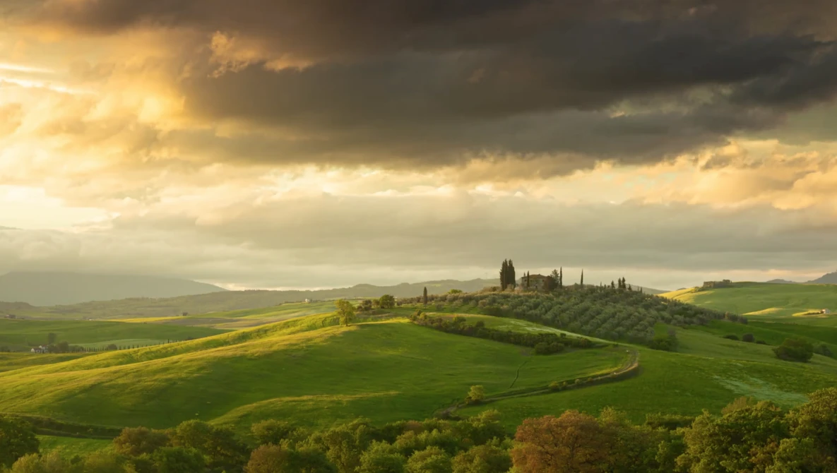 Rolling Tuscan hills with golden wheat fields, cypress trees, and dramatic storm clouds at sunset.