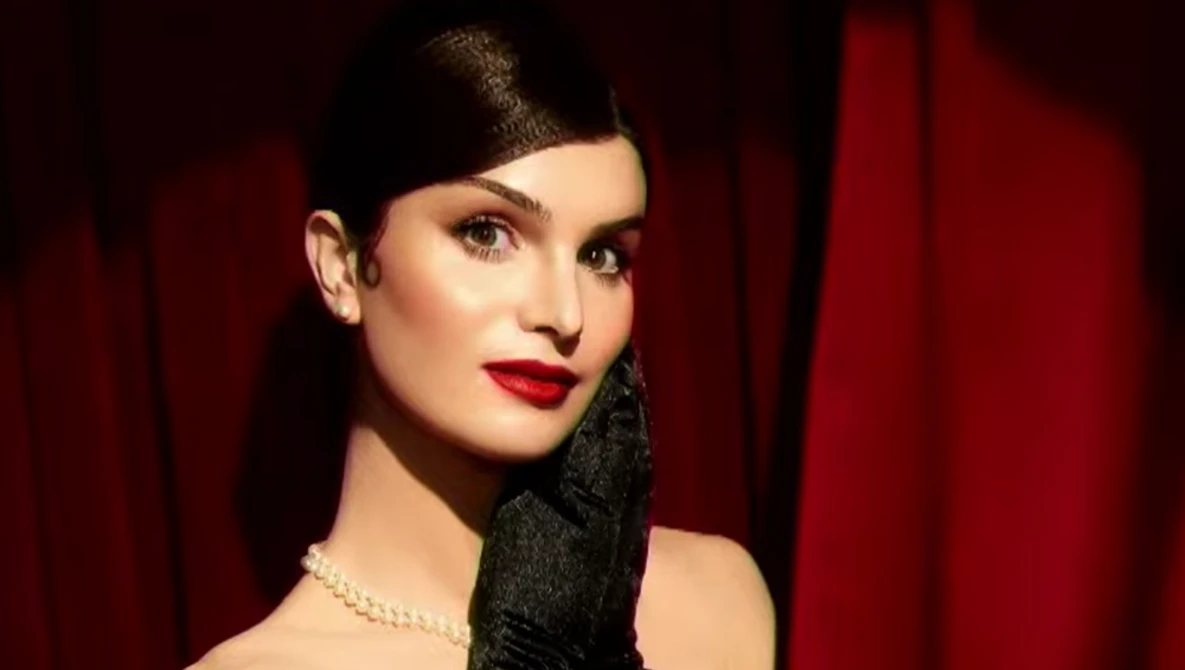 Woman in profile wearing red lipstick and black gloves against dark red curtain backdrop.