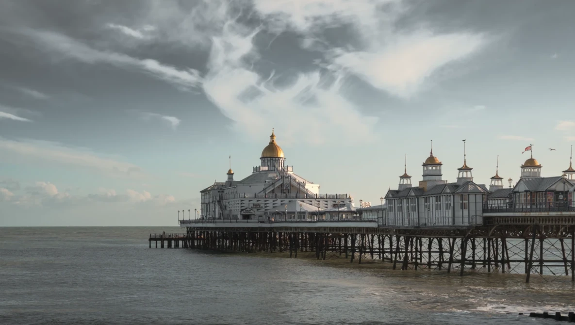 Historic seaside pier with ornate Victorian architecture and golden domes extending into calm water under dramatic cloudy sky.