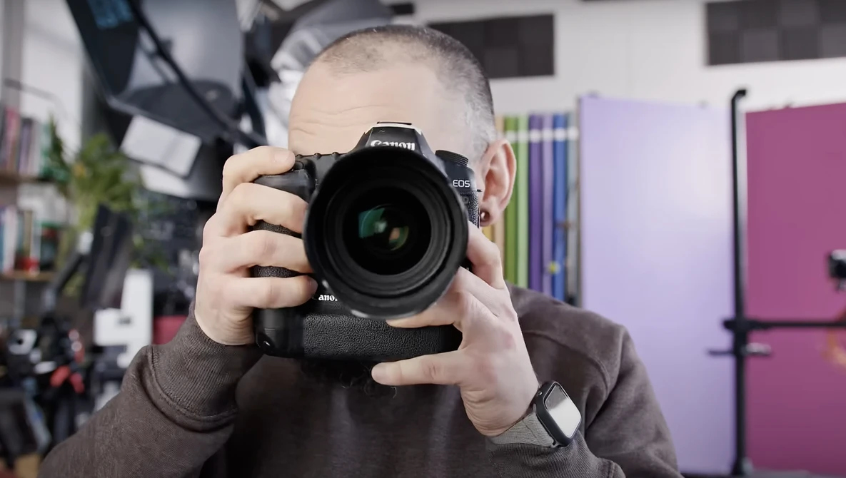 Young photographer holding a DSLR camera with battery grip up to his face in a studio setting.
