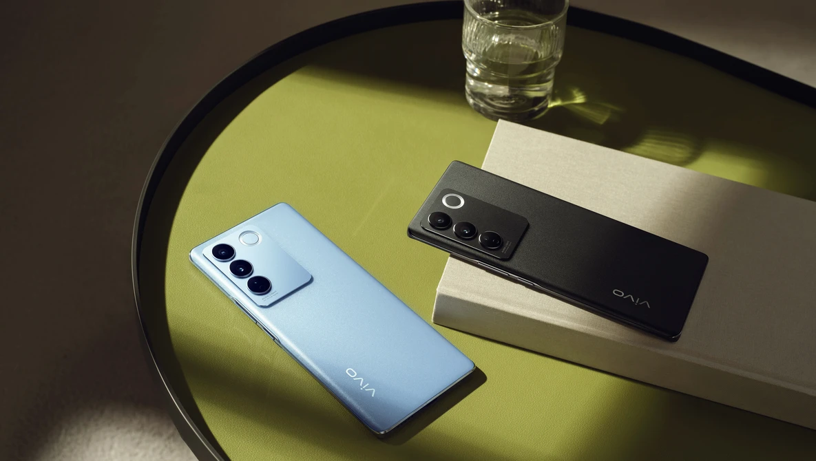 Two smartphones with triple rear camera setups displayed on a yellow-green surface next to a glass of water.