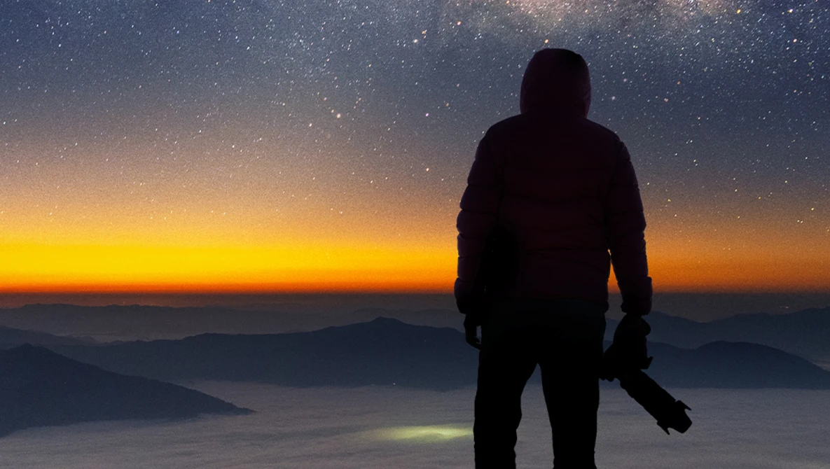 Silhouetted figure standing on a mountain ridge at sunrise, gazing toward a starry sky and golden horizon above layered clouds.