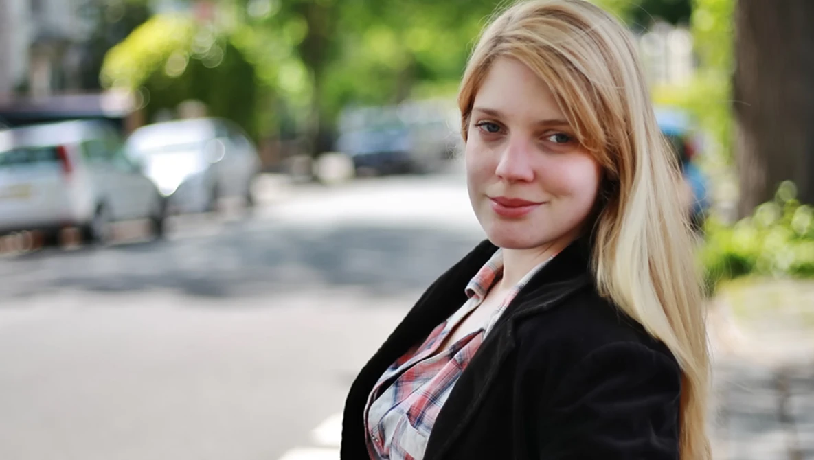 Woman with blonde hair wearing a black blazer and plaid shirt, smiling at camera outdoors.