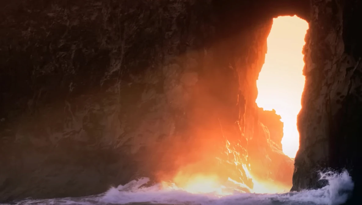 Brilliant sunlight streams through a narrow cave opening onto crashing waves and rocky shores.