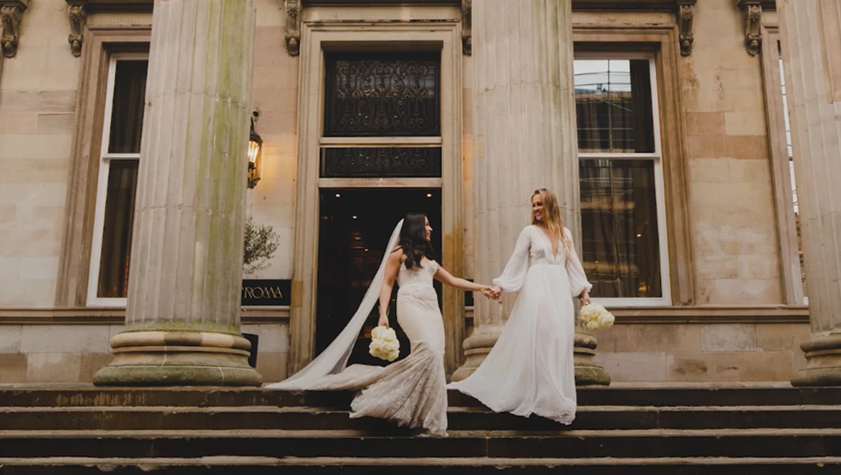 Two brides in flowing white wedding gowns with dramatic trains pose on classical stone steps holding bouquets.