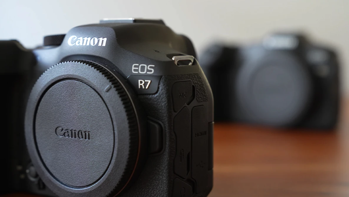 Canon EOS R7 mirrorless camera body displayed from front in sharp focus with additional camera equipment blurred in the background.
