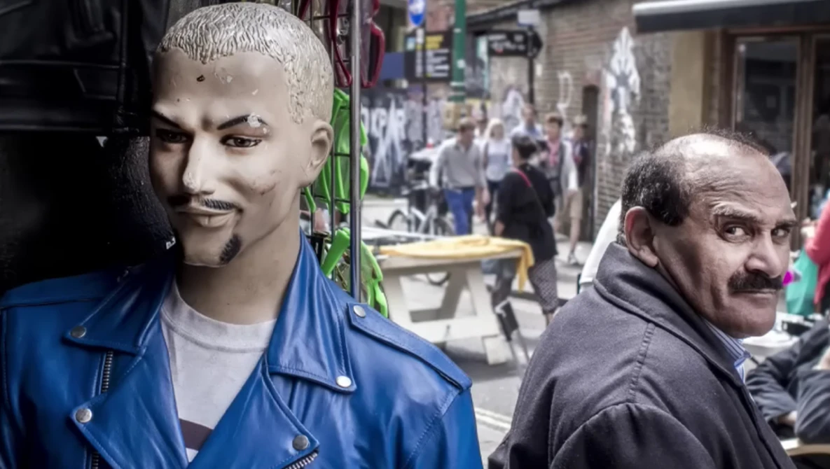 Man with mustache looking over shoulder at camera, street scene with mannequin head in blue jacket visible in background.