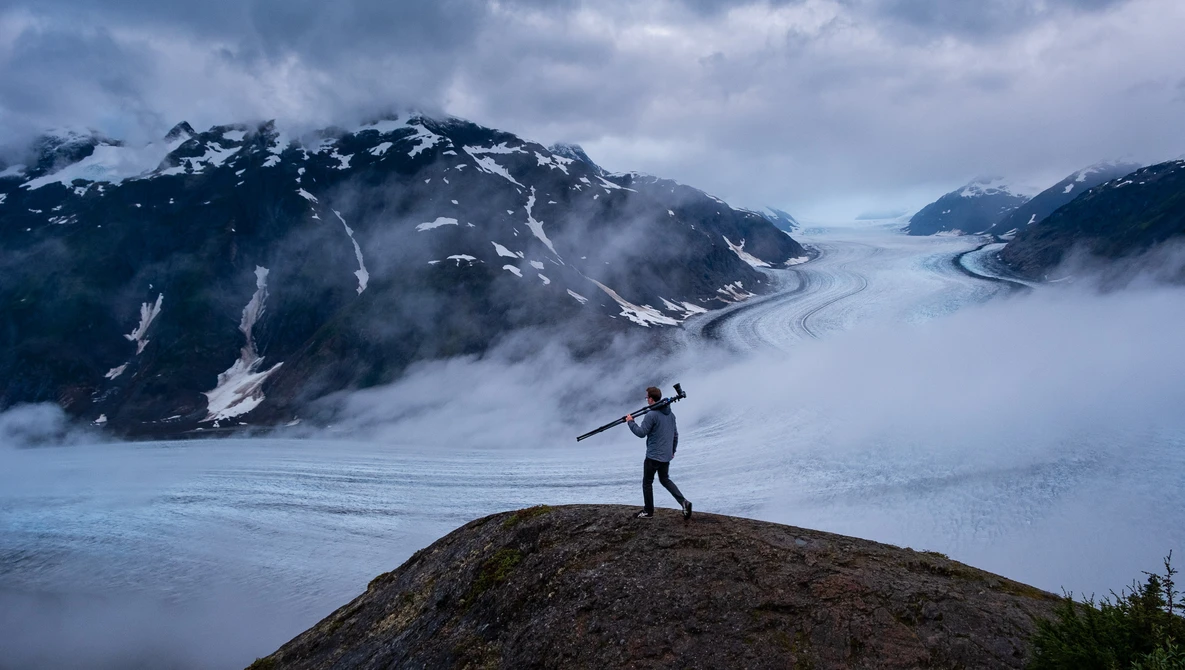 Hiker standing on a rocky outcrop overlooking a vast glacier valley surrounded by snow-capped mountains.