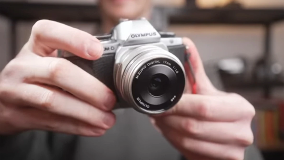 Hands holding an Olympus mirrorless camera with a prime lens attached.