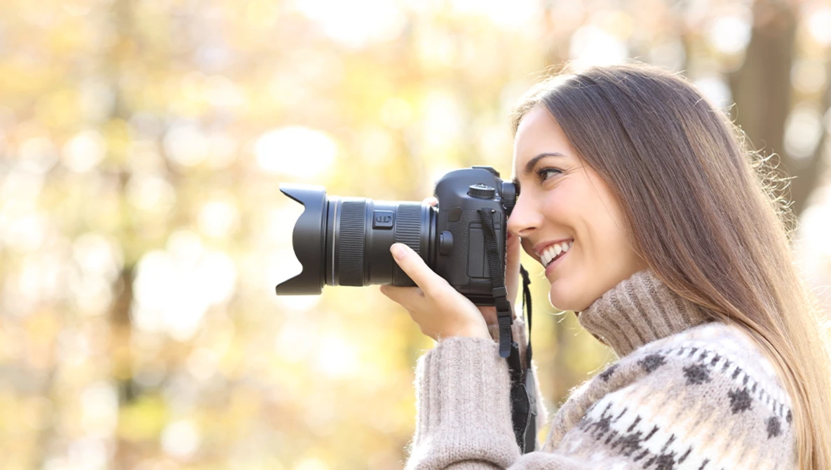 Woman holding a DSLR camera with telephoto lens, photographing outdoors with warm backlighting.
