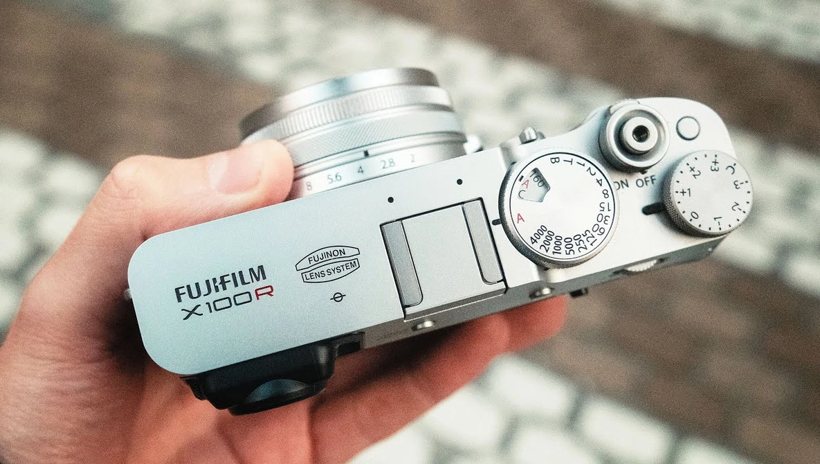White Fujifilm X100R rangefinder camera held in a hand, showing its manual controls and silver lens.