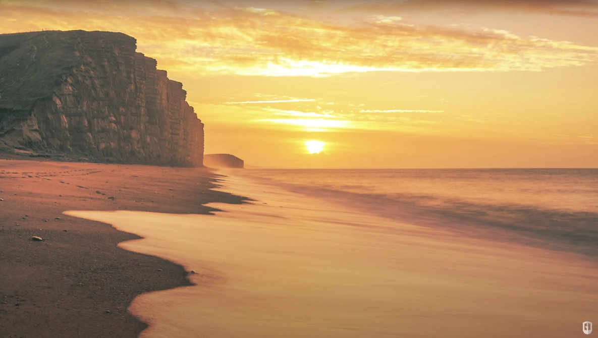 Golden sunrise over a beach with dramatic cliff formations silhouetted against the sky.