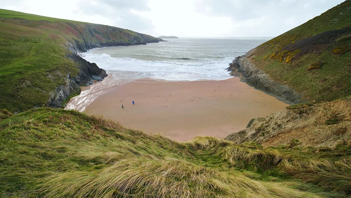 Secluded sandy beach nestled between green coastal cliffs with breaking waves and two distant figures.